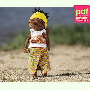 Könnte beinhalten: Häkelpuppen-Anleitung als PDF zum Download. Die Puppe ist braun mit einem gelben Kopftuch, einem weißen Oberteil und einem bunten Rock. Die Puppe steht auf einem Sandstrand.
