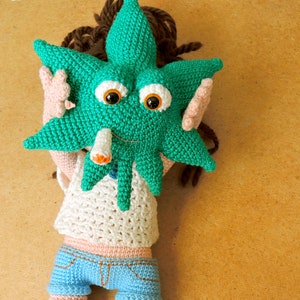 May include: A crocheted doll wearing a white shirt and blue jeans. The doll has a green marijuana leaf for a head with googly eyes and a cigarette.
