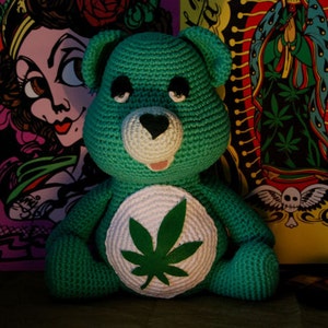 May include: A green crocheted teddy bear with a white patch on its chest featuring a green marijuana leaf. The bear is sitting in front of a colorful background with various designs.