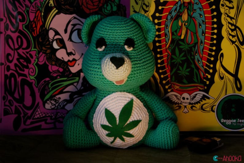 May include: A green crocheted teddy bear with a white patch on its chest featuring a green marijuana leaf. The bear is sitting in front of a colorful background with various designs.