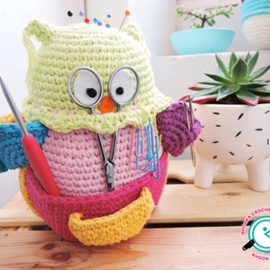 May include: A crocheted owl-shaped pincushion with a pink body, yellow beak, and green head. The owl is holding a pair of scissors, a crochet hook, and a red knitting needle. The owl is sitting on a wooden surface.