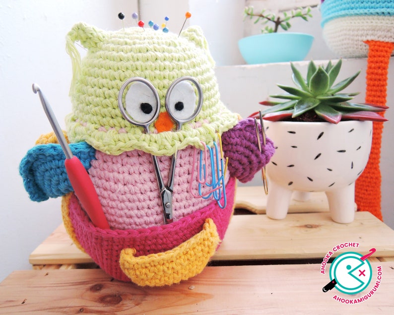 May include: A crocheted owl-shaped pincushion with a pink body, yellow beak, and green head. The owl is holding a pair of scissors, a crochet hook, and a red knitting needle. The owl is sitting on a wooden surface.
