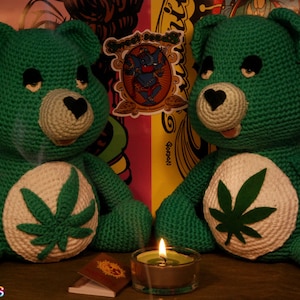 May include: Two green crocheted Care Bears with marijuana leaf designs on their chests. The bears are sitting on a wooden surface with a lit candle between them.