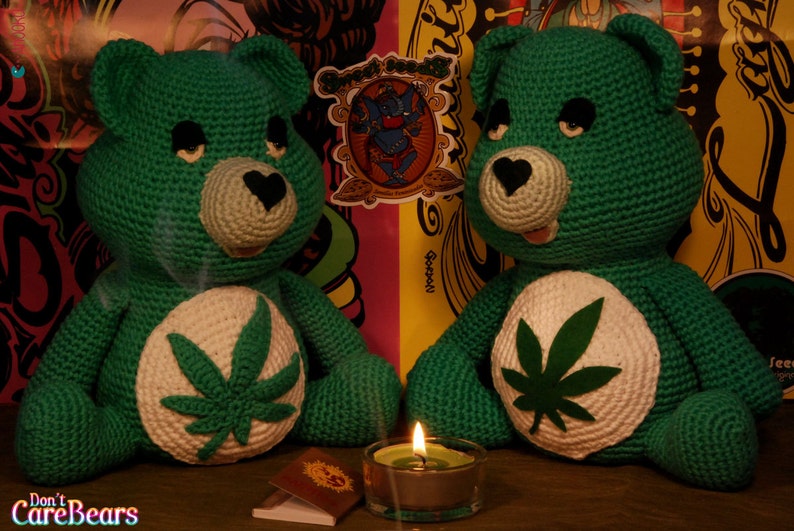 May include: Two green crocheted Care Bears with marijuana leaf designs on their chests. The bears are sitting on a wooden surface with a lit candle between them.