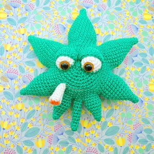 May include: A green crocheted marijuana leaf with googly eyes and a white cigarette in its mouth. The leaf is on a light blue background with a floral pattern.