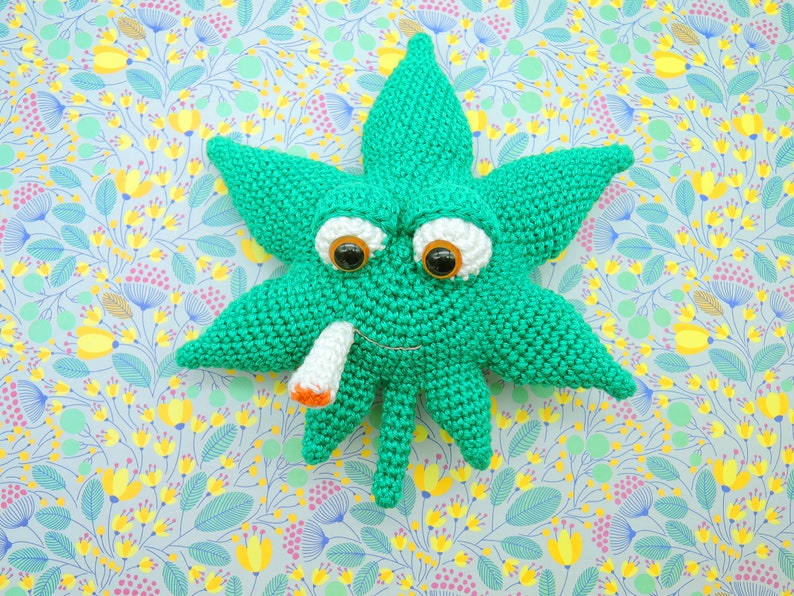 May include: A green crocheted marijuana leaf with googly eyes and a white cigarette in its mouth. The leaf is on a light blue background with a floral pattern.