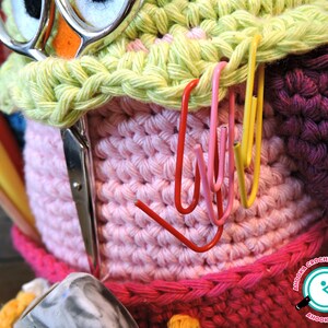 May include: A pink and green crocheted owl-shaped yarn holder with a pair of scissors and various coloured yarn needles sticking out of the top. The owl has a pair of googly eyes and a pink beak. The yarn holder is decorated with colourful paper clips.