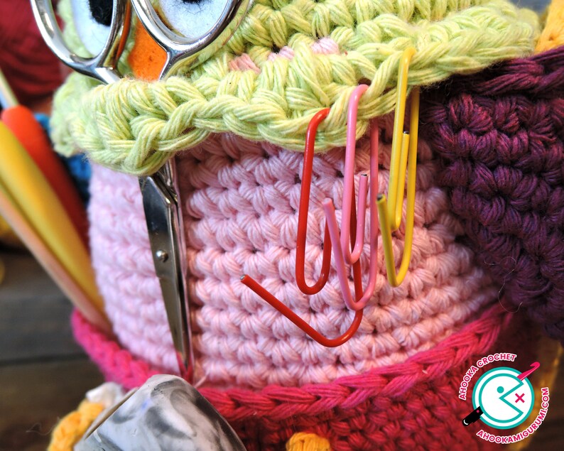 May include: A pink and green crocheted owl-shaped yarn holder with a pair of scissors and various coloured yarn needles sticking out of the top. The owl has a pair of googly eyes and a pink beak. The yarn holder is decorated with colourful paper clips.