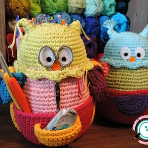 May include: Two crocheted owl-shaped containers, one in pink, green, and yellow with a variety of sewing needles and tools inside, and the other in blue, green, and purple. Both owls have large black eyes and a small orange beak.