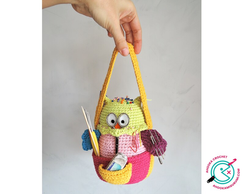 May include: A crocheted owl-shaped bag with a yellow handle. The bag is filled with colourful yarn, needles, and pins. The owl has large, round eyes and a pink beak.