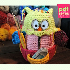Peut inclure: Un porte-fil en forme de hibou crocheté avec un corps rose, une tête verte et un bec jaune. Le hibou a de grands yeux noirs et tient divers aiguilles à tricoter et crochets. Le hibou est assis sur une surface en bois.