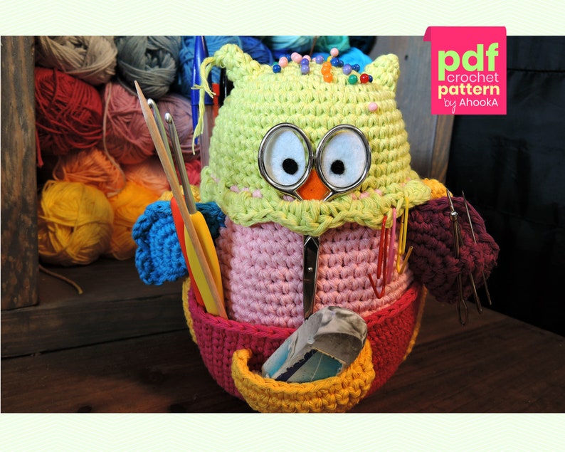 May include: A crocheted owl-shaped yarn holder with a pink body, green head, and yellow beak. The owl has large black eyes and is holding various yarn needles and crochet hooks. The owl is sitting on a wooden surface.