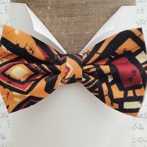 May include: A bow tie with a geometric pattern in shades of orange, black, red, and white. The bow tie is made of fabric and has a textured surface.
