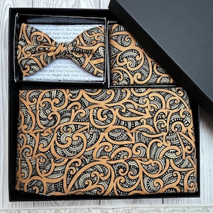 Puede incluir: Un conjunto de ropa formal en una caja negra, que incluye una pajarita, un pañuelo de bolsillo y un fajín. El conjunto presenta un patrón de remolino negro y bronceado que se repite. La caja está abierta, revelando el contenido. El texto "Dapper Jack Hand Crafted Bow Ties Cravats Accessories" es visible.