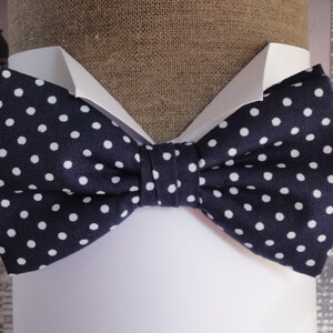 May include: A navy blue bow tie with white polka dots. The bow tie is made of fabric and has a classic shape.