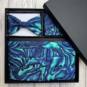 May include: A formal set in a black box, including a bow tie, pocket square, and cummerbund. The items display a blue and teal marbled design. The bow tie is pre-tied. The cummerbund is folded and ready to wear.