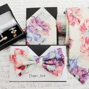 May include: A set of men's accessories featuring a floral patterned bow tie, pocket square, and tie. The accessories are in shades of pink, purple, and white. The set also includes a box of silver cufflinks.