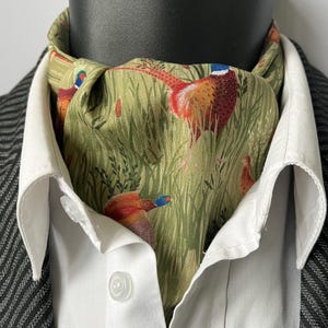 Puede incluir: Un pañuelo de cuello verde con estampado de faisanes, usado con una camisa blanca con cuello y un blazer de espiga gris oscuro. El pañuelo de cuello presenta coloridos faisanes en un diseño de campo herboso.