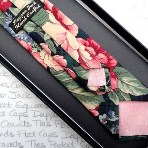 May include: A dark blue tie with a floral pattern in shades of pink, green, and cream. The tie is hand-crafted and comes with a matching pocket square. The label reads "Dapper Jack Hand Crafted".