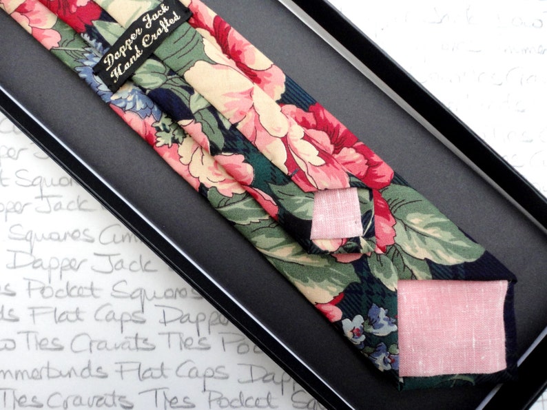 May include: A dark blue tie with a floral pattern in shades of pink, green, and cream. The tie is hand-crafted and comes with a matching pocket square. The label reads "Dapper Jack Hand Crafted".