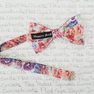 May include: A floral patterned bow tie with a white background and pink, purple, and blue flowers. The bow tie is attached to a matching patterned strap. The bow tie has a label that says "Dapper Jack".