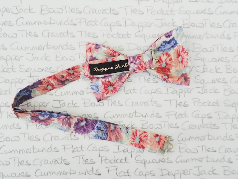 May include: A floral patterned bow tie with a white background and pink, purple, and blue flowers. The bow tie is attached to a matching patterned strap. The bow tie has a label that says "Dapper Jack".