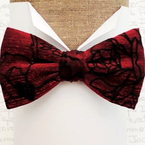 May include: A red bow tie with a black floral pattern. The bow tie is made of a silky fabric and has a slightly textured surface.