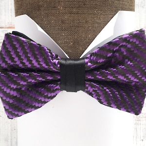 May include: A purple and black bow tie with a carbon fiber pattern. The bow tie is made of a woven fabric and has a black satin band in the center.
