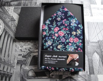 Può includere: Un fazzoletto da taschino blu scuro con un motivo floreale rosa, bianco e verde. Il fazzoletto da taschino è in una scatola nera con un'etichetta bianca che dice "Dapper Jack Hand Crafted Bow Ties, Cravats, Accessories www.dapperjack.co.uk mob: 07887898331"