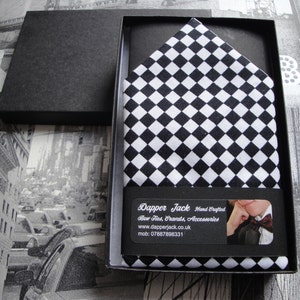 May include: A black and white checked pocket square in a black box. The box has a label that says "Dapper Jack Hand Crafted Bow Ties, Cravats, Accessories www.dapperjack.co.uk mob: 07887896331"