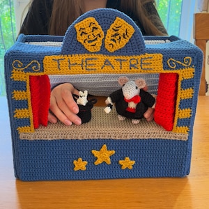 May include: A handmade blue crochet theater with the word "THEATRE" across the top. The theater features a stage with two small crochet mouse puppets. The top of the theater has two yellow masks. The theater is decorated with yellow stars.