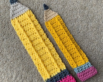 May include: Two crocheted pencil-shaped items, one larger than the other. The larger pencil is yellow with a black and pink eraser. The smaller pencil is yellow with a gray and pink eraser.