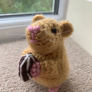 May include: A crocheted brown hamster holding a chocolate and white striped cookie. The hamster has pink ears, nose, and feet.