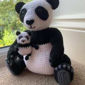 May include: A crocheted panda bear holding a smaller crocheted panda bear. Both pandas are white with black markings around their eyes and ears. The larger panda has black paws and a black belly.