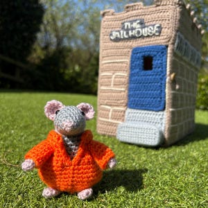 Può includere: Un topo all'uncinetto con una giacca arancione è in piedi davanti a una prigione all'uncinetto. Il topo è grigio con orecchie e piedi rosa. La prigione è beige con una porta blu e la scritta "THE JAILHOUSE" sopra.