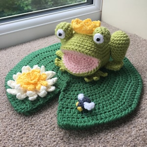 Puede incluir: Una rana verde de crochet con una corona amarilla se sienta en un nenúfar verde de crochet con una flor blanca y una abeja negra y amarilla de crochet.