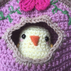 May include: A crocheted birdhouse with a pink flower on top and a yellow chick peeking out of the opening.