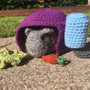 May include: A gray crocheted guinea pig wearing a purple crocheted hat, a blue crocheted hat, and a green crocheted carrot.