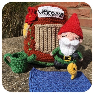 Może przedstawiać: Zestaw szydełkowych krasnali ogrodowych z czerwoną czapką, białą brodą i zielonym strojem. Zawiera miniaturowy domek z napisem "Welcome", zieloną konewkę, niebieską matę i żółtą rybę.