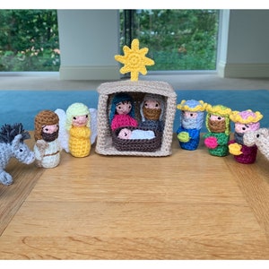 Puede incluir: Un belén tejido a ganchillo con figuras de María, José, el niño Jesús, ángeles, reyes magos, un burro, una oveja y un camello. Las figuras son de varios colores, incluyendo beige, azul, amarillo y rosa. Una estrella amarilla está encima del establo.