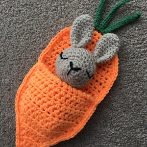 May include: A crocheted carrot-shaped stuffed toy with a small gray bunny inside. The carrot is orange and the bunny has black eyes and a closed mouth.