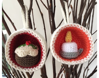 May include: Two crocheted Christmas ornaments hanging on a branch. One ornament is a red and white crocheted cup with a brown and white crocheted Christmas pudding inside. The other ornament is a red and white crocheted cup with a white crocheted candle inside.