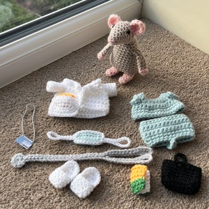 Op de afbeelding: Een gehaakte grijze muis met roze oren die een witte gehaakte doktersjas, een lichtblauwe gehaakte outfit, een wit gehaakt masker, een witte gehaakte ketting, een paar witte gehaakte schoenen, een gehaakte gele en groene sushirol en een zwarte gehaakte tas draagt.