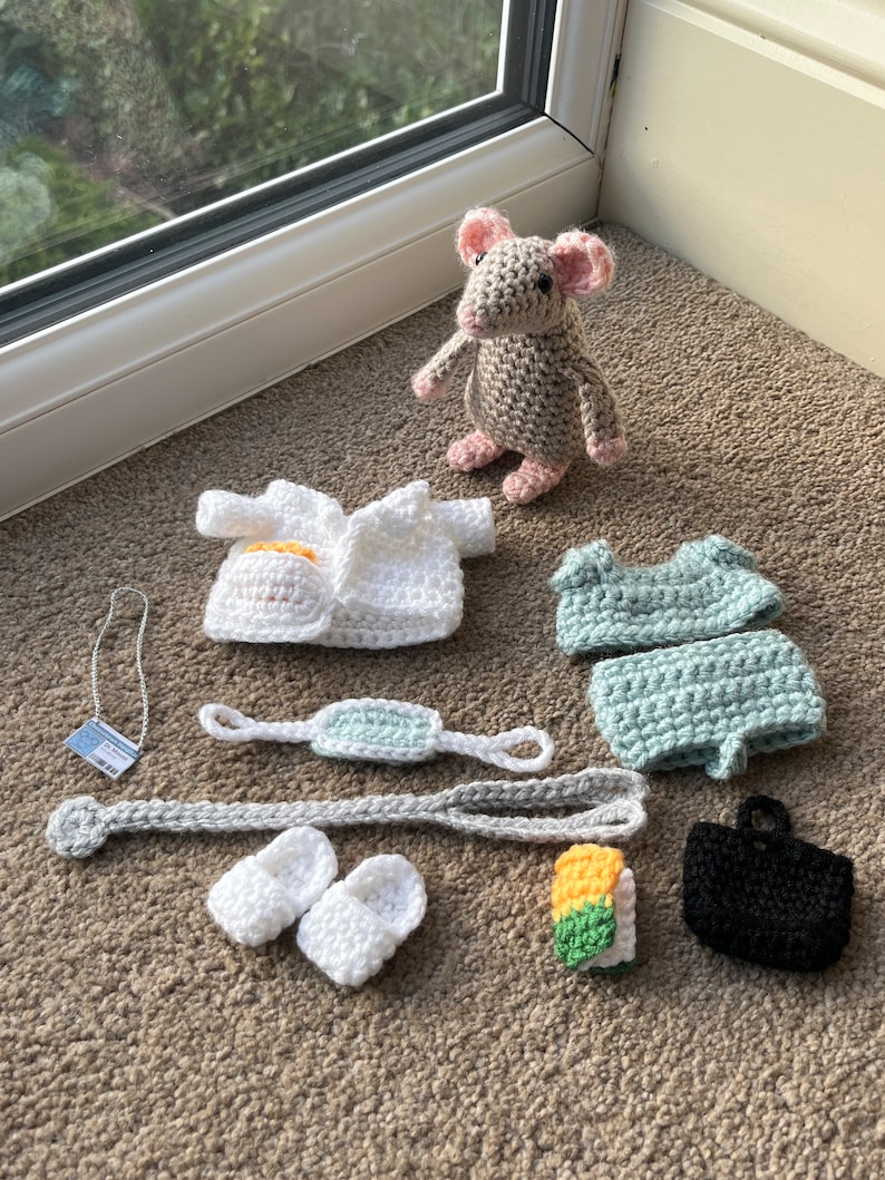 Op de afbeelding: Een gehaakte grijze muis met roze oren die een witte gehaakte doktersjas, een lichtblauwe gehaakte outfit, een wit gehaakt masker, een witte gehaakte ketting, een paar witte gehaakte schoenen, een gehaakte gele en groene sushirol en een zwarte gehaakte tas draagt.