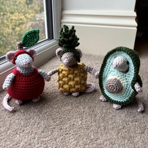 May include: Three crocheted mice dressed as fruits: a red apple, a yellow pineapple, and a green avocado. Each mouse has gray bodies, pink ears, and small tails. The apple mouse wears a red dress and a green leaf. The pineapple mouse wears a yellow dress. The avocado mouse is inside a green and blue avocado.