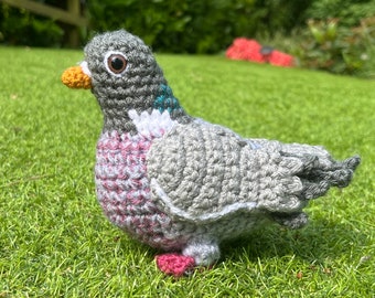 May include: A crocheted grey and pink pigeon with a teal eye and orange beak. The pigeon is standing on green grass.