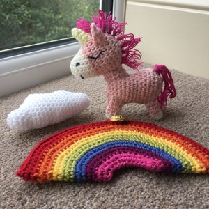 May include: A pink crocheted unicorn with a yellow horn and pink mane standing on a rainbow crocheted rug with a white crocheted cloud.