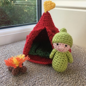 May include: A handmade crochet camping set featuring a red tent with a yellow flag, a small campfire, and a green gnome-like figure. The tent has a green interior, and the campfire has orange and yellow flames.