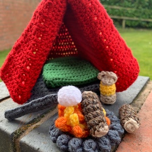Pu&ograve; includere: Una tenda rossa all'uncinetto con un sacco a pelo verde all'interno. Un fal&ograve; all'uncinetto con tronchi e un marshmallow bianco in cima si trova davanti alla tenda.
