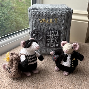 Peut inclure: Coffre-fort gris crocheté avec le mot "VAULT" brodé en or. Trois souris crochetées sont devant le coffre-fort, l'une portant une tenue noire et blanche, une en costume et une avec un petit sac.